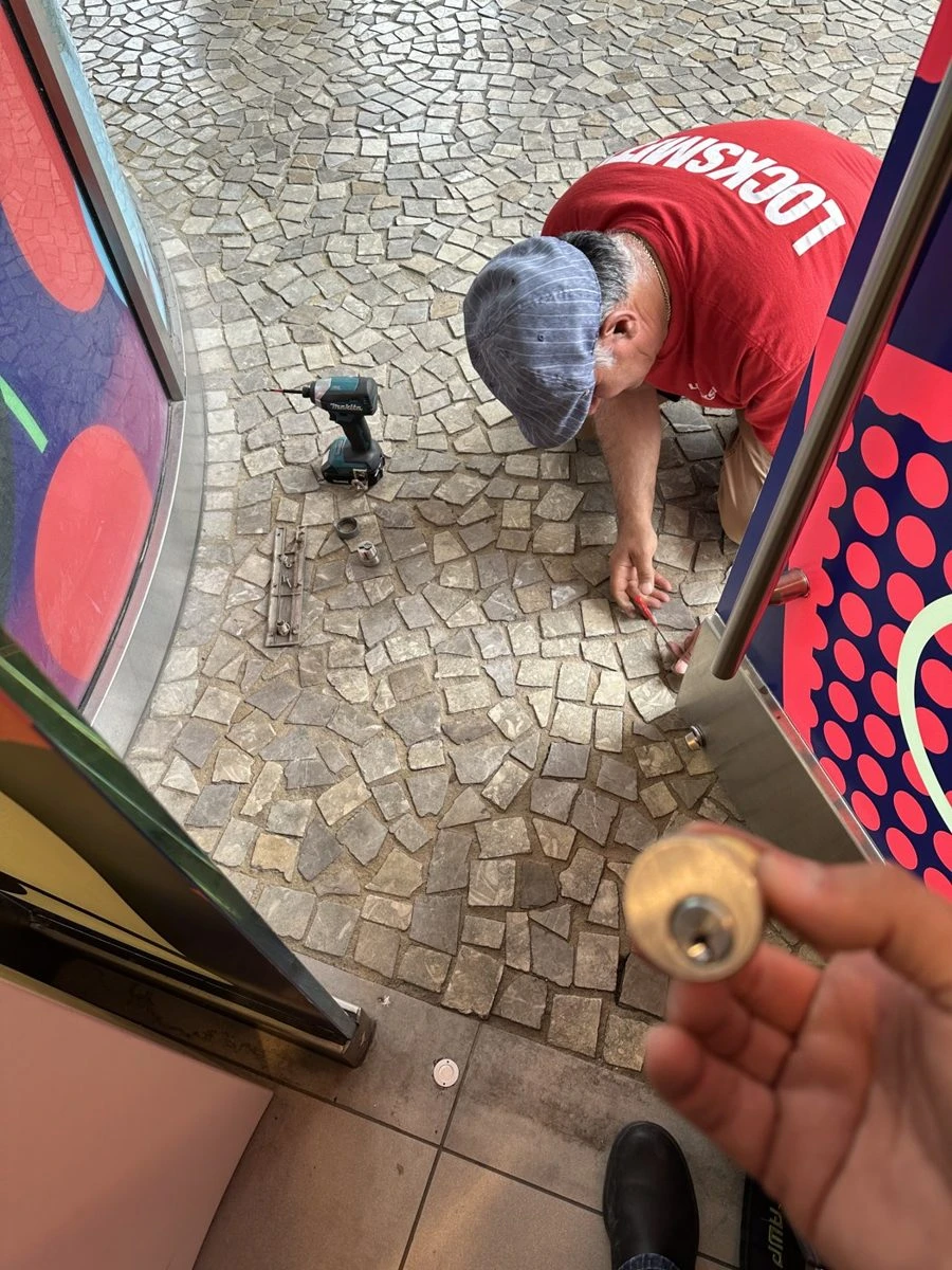 Locksmith holds a pulled lock cylinder toward the camera while a second tech in a red LOCKSMITH shirt works on a storefront door frame