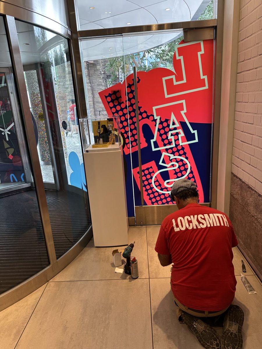 Locksmith kneeling at a glass storefront door with a drill and parts on the floor, LOCKSMITH printed on his shirt