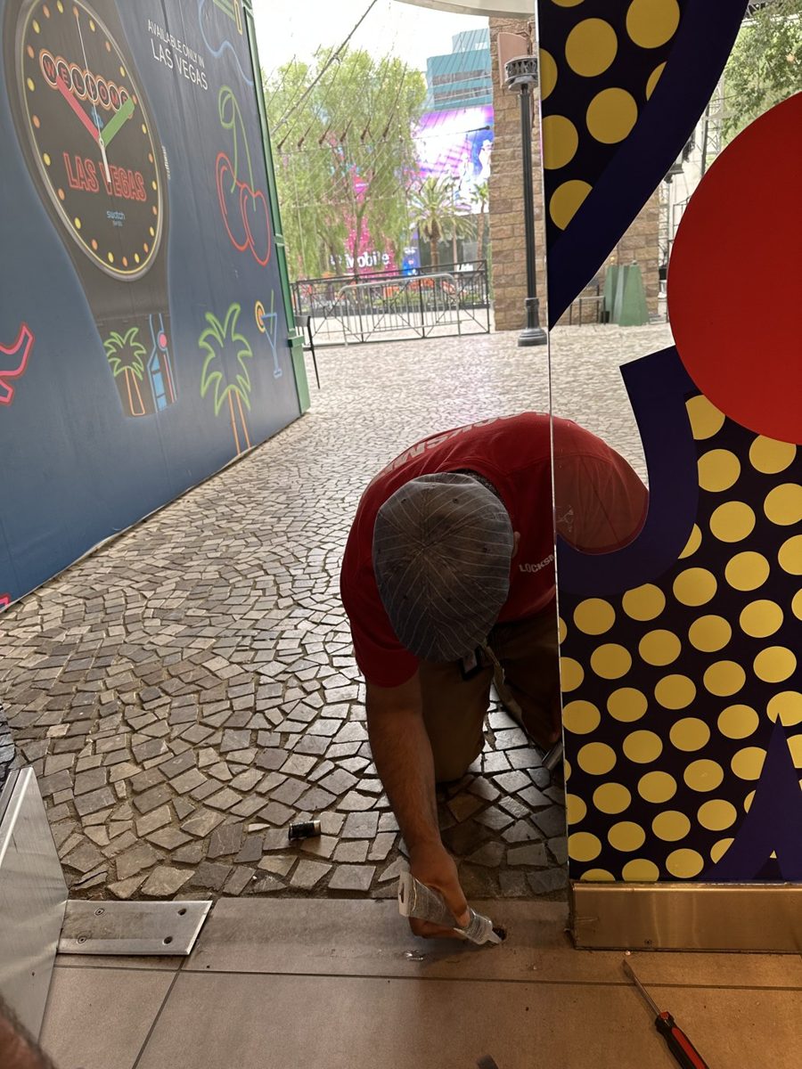 Locksmith in a red shirt kneels at a storefront threshold on the Las Vegas Strip, working on the door base with a hand tool